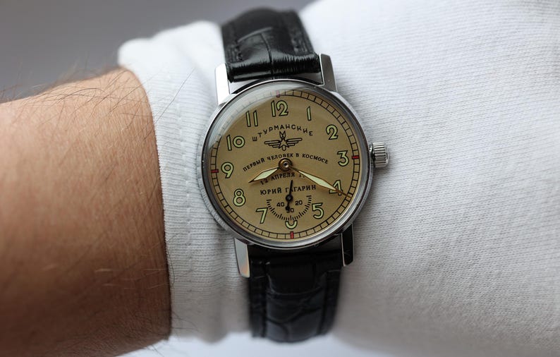 May include: A vintage wristwatch with a silver case and a tan face. The watch features black numerals, hands, and a black leather band. The watch face has Cyrillic text and the name "Yuri Gagarin".