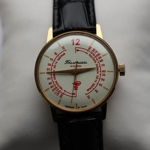 May include: A vintage wristwatch with a gold-toned case and a black leather strap. The watch face is cream-colored with red numerals and markings, including the number 12 in red. The watch has a small crown on the side.