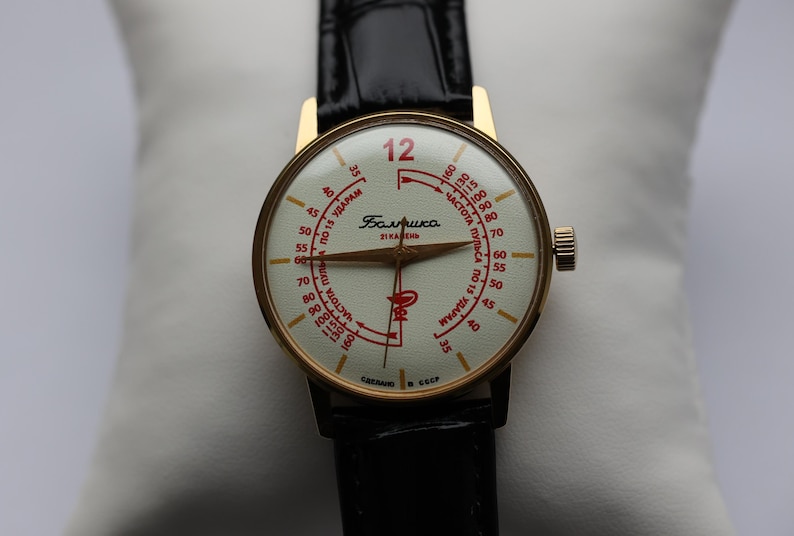 May include: A vintage wristwatch with a gold-toned case and a black leather strap. The watch face is cream-colored with red numerals and markings, including the number 12 in red. The watch has a small crown on the side.