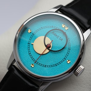 May include: A silver wristwatch with a turquoise blue face and gold accents. The watch face has a smaller circle with the word "PAKEITA" in black lettering.