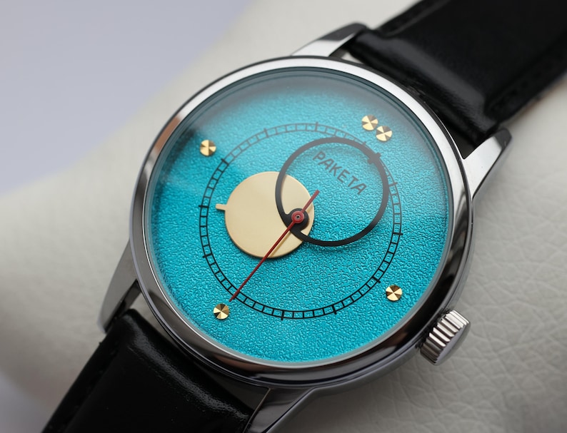 May include: A silver wristwatch with a turquoise blue face and gold accents. The watch face has a smaller circle with the word "PAKEITA" in black lettering.