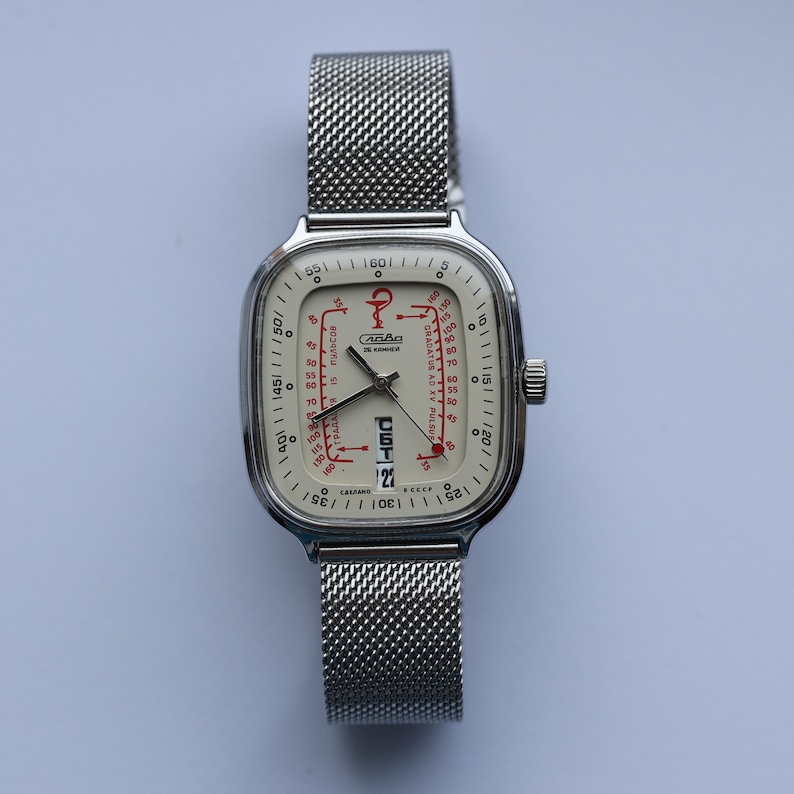 May include: A vintage wristwatch with a square, silver-toned case and a mesh band. The watch face is off-white with red and black markings, including the date. The watch has a small crown on the side and is set against a light background.