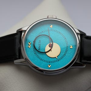 May include: A silver wristwatch with a blue face and a black leather band. The face has a white circle in the center and the word "RAKETTA" printed in black.