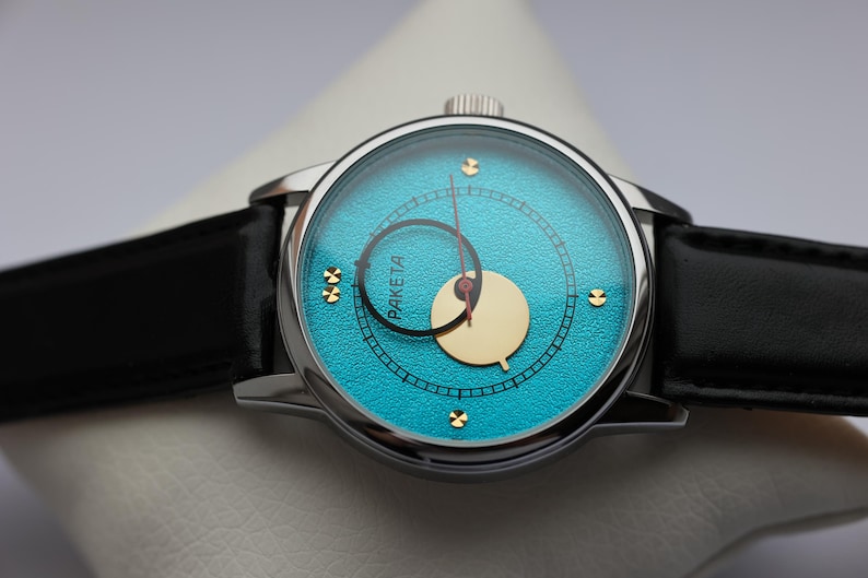 May include: A silver wristwatch with a blue face and a black leather band. The face has a white circle in the center and the word "RAKETTA" printed in black.
