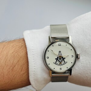 May include: A silver watch with a white face and a black Masonic symbol. The watch has a silver mesh band.