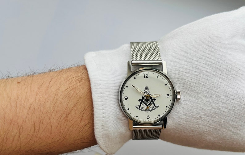 May include: A silver watch with a white face and a black Masonic symbol. The watch has a silver mesh band.