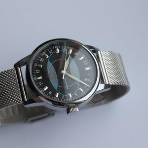 May include: A silver wristwatch with a black face featuring blue and red geometric designs. The watch has a mesh band and a silver case. The watch face displays the numbers 1 through 24, and the word "ПОЛЯРНЫЕ" is visible.
