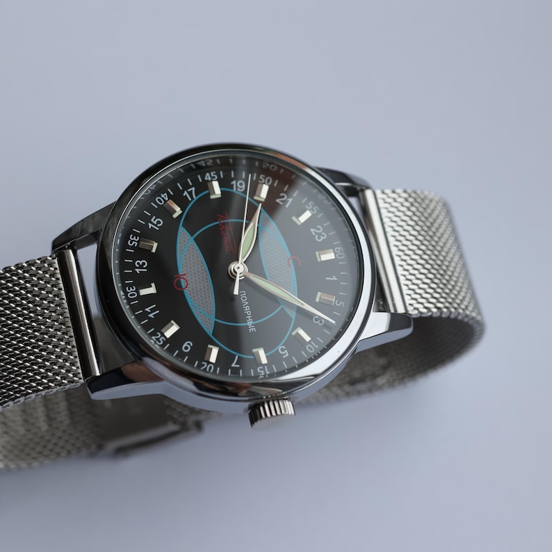 May include: A silver wristwatch with a black face featuring blue and red geometric designs. The watch has a mesh band and a silver case. The watch face displays the numbers 1 through 24, and the word "ПОЛЯРНЫЕ" is visible.