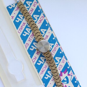 May include: A gold-toned wristwatch with a round face and a metal band. The watch is on a blue and white box with the text "ЧАКА" repeated in different sizes and orientations.
