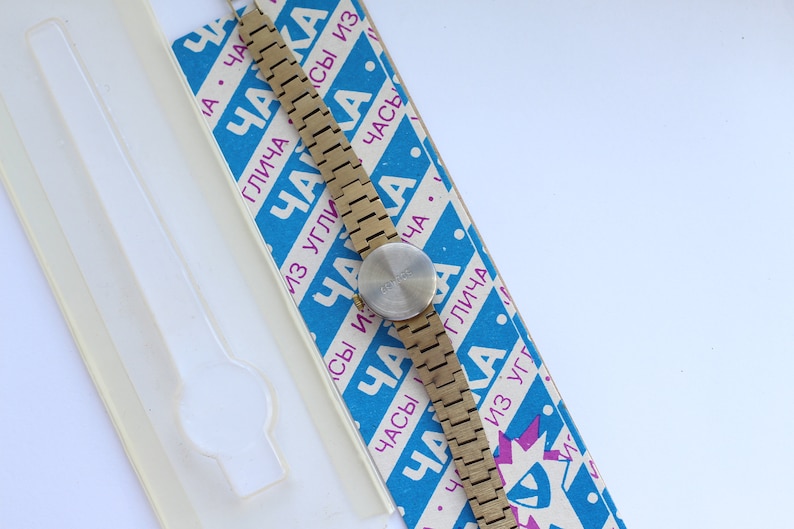 May include: A gold-toned wristwatch with a round face and a metal band. The watch is on a blue and white box with the text "ЧАКА" repeated in different sizes and orientations.