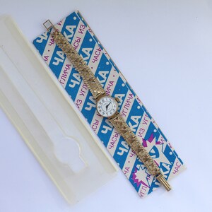 May include: A gold-tone wristwatch with a white face and black Roman numeral hour markers. The watch is on a gold-tone metal band with a textured pattern. The watch is on a blue and white box with the text "Чака" in pink and blue.