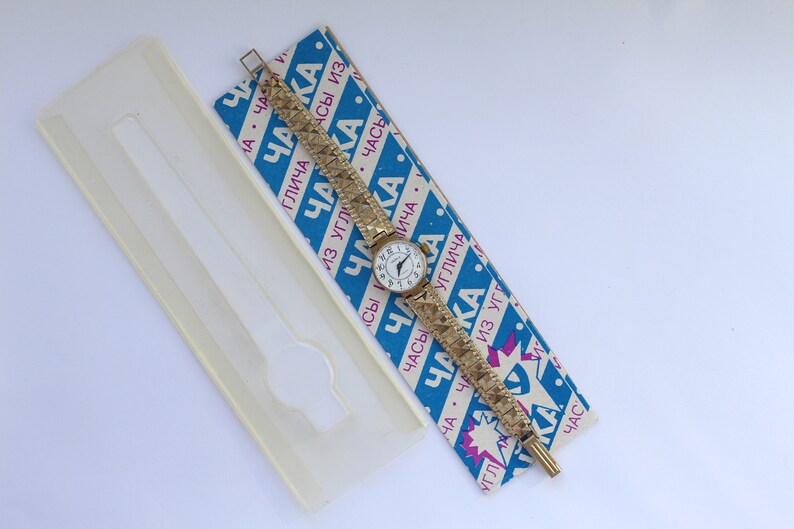 May include: A gold-tone wristwatch with a white face and black Roman numeral hour markers. The watch is on a gold-tone metal band with a textured pattern. The watch is on a blue and white box with the text "Чака" in pink and blue.