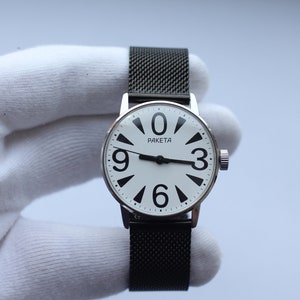 May include: A silver wristwatch with a white face and black numbers. The watch has a black mesh strap and says "PAKEta" on the face.