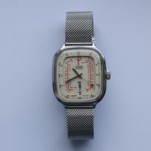 May include: A vintage wristwatch with a square, silver-toned case and a mesh band. The off-white face features red and black markings, including the date and a unique graphic design. The watch has a small crown on the side and is set against a light background.
