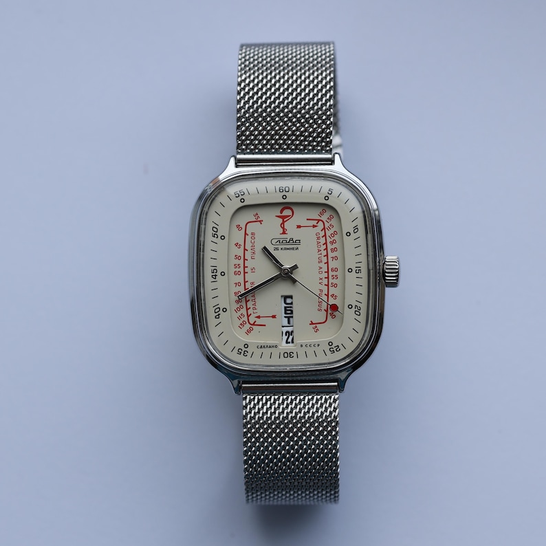 May include: A vintage wristwatch with a square, silver-toned case and a mesh band. The off-white face features red and black markings, including the date and a unique graphic design. The watch has a small crown on the side and is set against a light background.