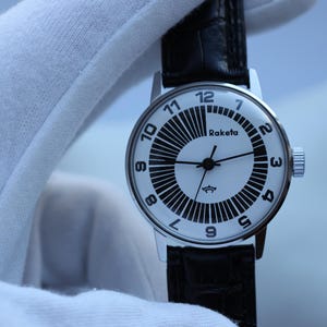 Könnte beinhalten: Eine silberne Armbanduhr mit weißem Zifferblatt und schwarzen Ziffern und Markierungen. Auf dem Zifferblatt steht das Wort "Raketa". Die Uhr hat ein schwarzes Lederarmband.