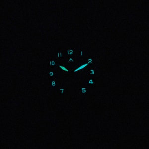 May include: A black watch face with luminous green hands and hour markers. The watch face has numbers 1 through 12 around the edge.