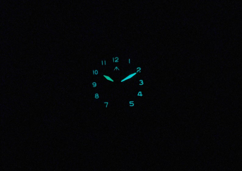May include: A black watch face with luminous green hands and hour markers. The watch face has numbers 1 through 12 around the edge.