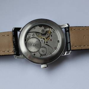 May include: A silver-toned wristwatch with a transparent back, showcasing the intricate internal gears and mechanisms. The watch has a black leather strap with a beige underside. The words "GENUINE LEATHER" are visible on the strap.