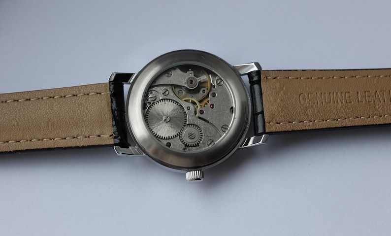 May include: A silver-toned wristwatch with a transparent back, showcasing the intricate internal gears and mechanisms. The watch has a black leather strap with a beige underside. The words "GENUINE LEATHER" are visible on the strap.