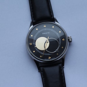 Puede incluir: Un reloj de pulsera plateado con una esfera negra y una correa de cuero negra. La esfera del reloj presenta una complicación de fase lunar con una luna de color dorado y un fondo negro.