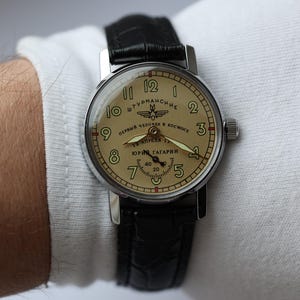 May include: A vintage wristwatch with a cream-colored face and black leather band. The watch face features black numerals, hands, and text in Cyrillic script. The watch is a Sturmanskie model, commemorating Yuri Gagarin's spaceflight.