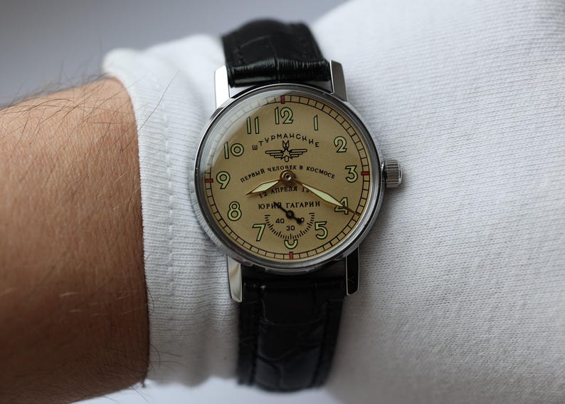 May include: A vintage wristwatch with a cream-colored face and black leather band. The watch face features black numerals, hands, and text in Cyrillic script. The watch is a Sturmanskie model, commemorating Yuri Gagarin's spaceflight.