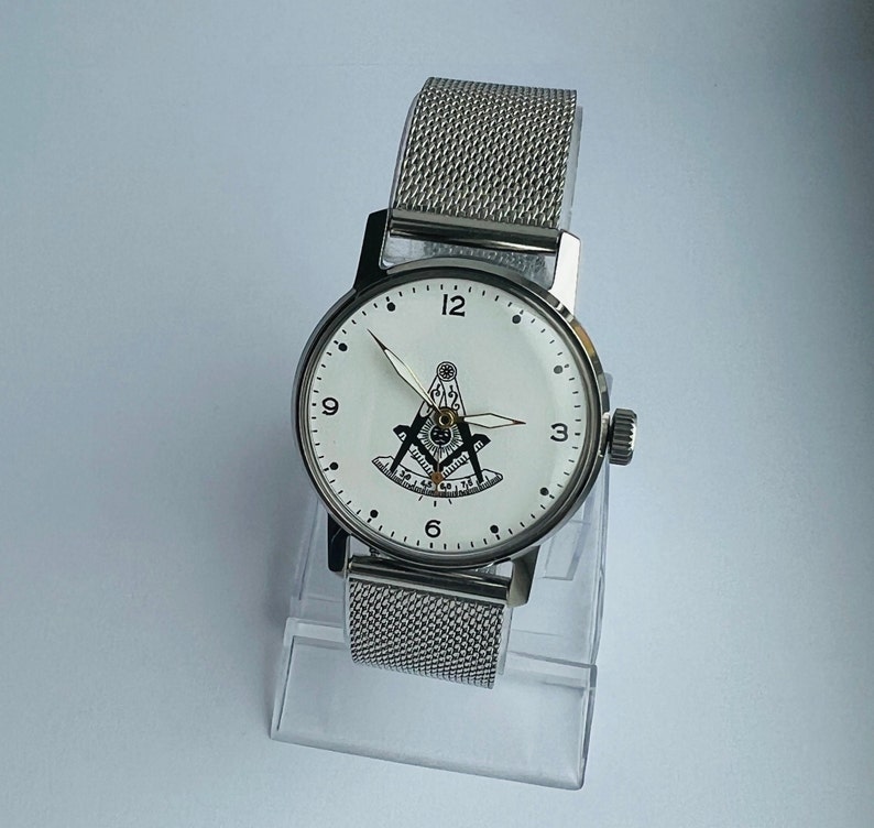 May include: A silver watch with a white face and a silver mesh band. The face of the watch features a black and white compass and the numbers 12, 3, 6, and 9.