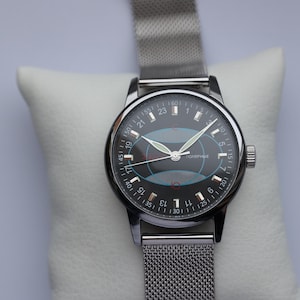 May include: A vintage wristwatch with a black face and silver-tone case and mesh band. The watch face features white numerals, green hands, and a blue and red graphic. The watch is displayed on a white cushion.