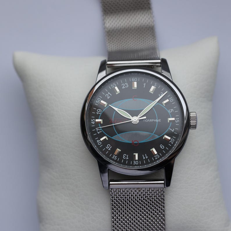 May include: A vintage wristwatch with a black face and silver-tone case and mesh band. The watch face features white numerals, green hands, and a blue and red graphic. The watch is displayed on a white cushion.