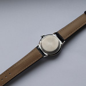 Puede incluir: Un reloj de pulsera plateado con una correa de cuero negro. La correa est&aacute; etiquetada como "Genuine Leather" y tiene una hebilla plateada.