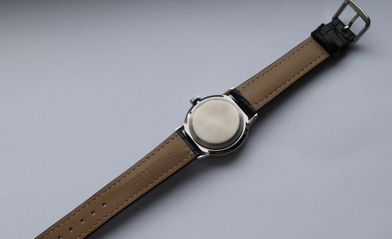 Puede incluir: Un reloj de pulsera plateado con una correa de cuero negro. La correa est&aacute; etiquetada como "Genuine Leather" y tiene una hebilla plateada.