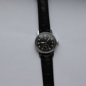 May include: A black-faced wristwatch with silver hands and numerals on a black leather band.