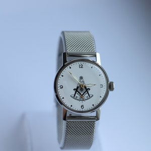 May include: A silver wristwatch with a white face and black markings. The watch face features a black and white compass and square design. The watch has a silver mesh band.