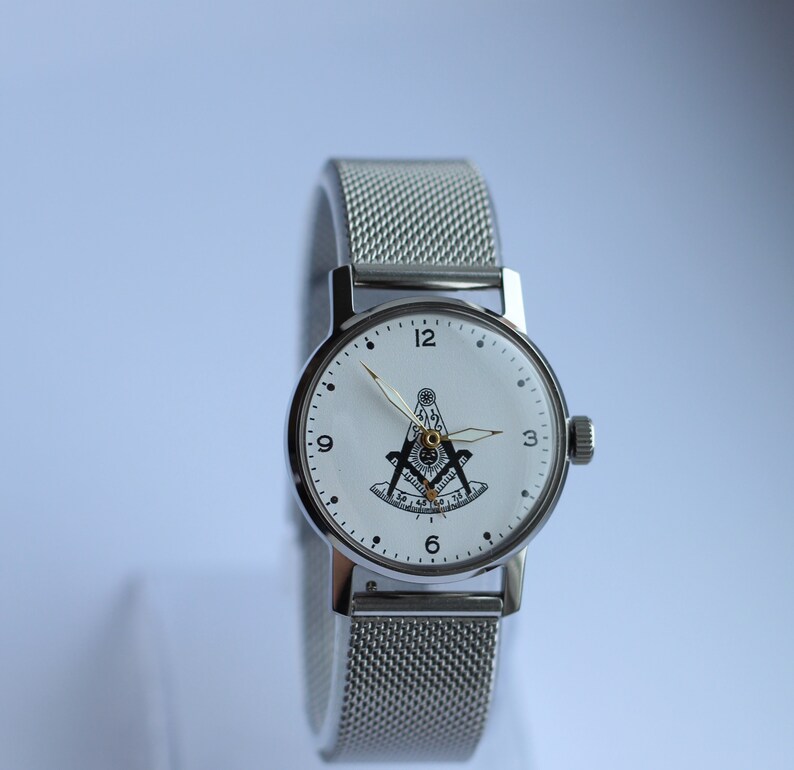 May include: A silver wristwatch with a white face and black markings. The watch face features a black and white compass and square design. The watch has a silver mesh band.