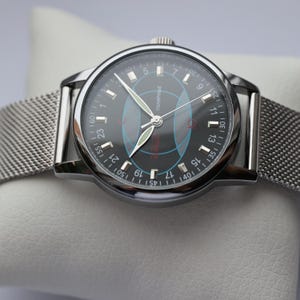 May include: A silver-toned wristwatch with a black face and a mesh band. The watch face features white numerals, a red logo, and blue and red geometric designs. The watch is displayed on a white surface.