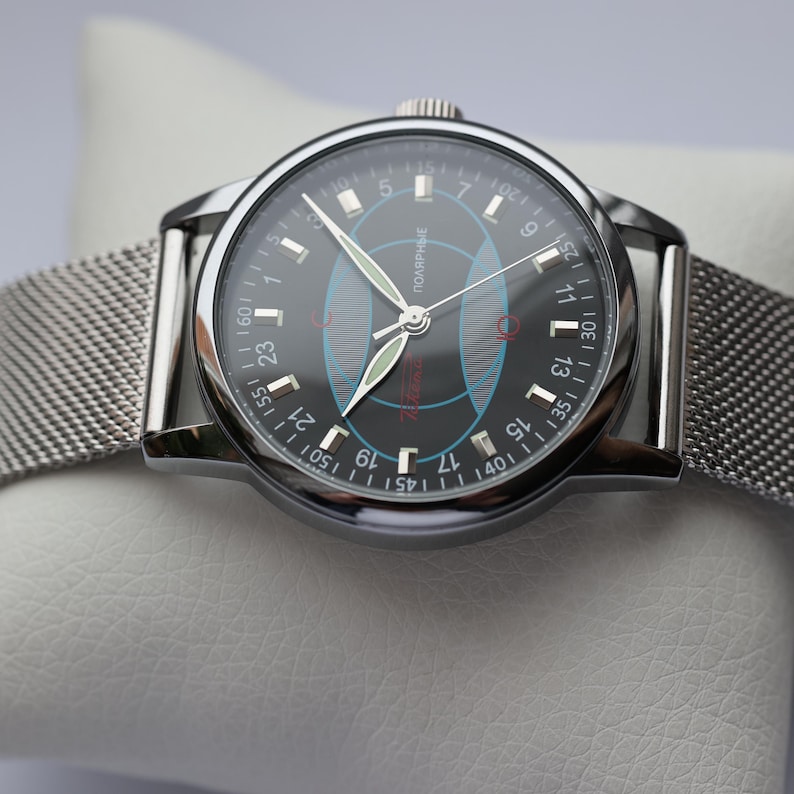 May include: A silver-toned wristwatch with a black face and a mesh band. The watch face features white numerals, a red logo, and blue and red geometric designs. The watch is displayed on a white surface.
