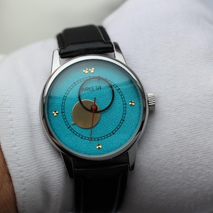 May include: A silver wristwatch with a blue face and gold accents. The watch has a black leather band and the words "PAKEJTA" printed on the face.