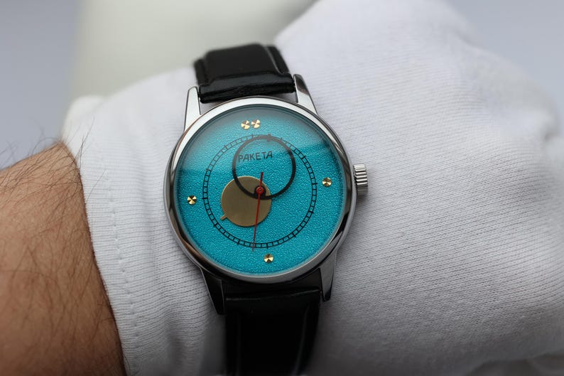 May include: A silver wristwatch with a blue face and gold accents. The watch has a black leather band and the words "PAKEJTA" printed on the face.