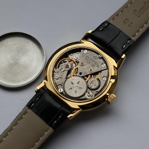 May include: A gold-toned wristwatch with a black leather strap, showcasing the intricate internal mechanisms. The watch face is open, revealing gears and components. The watch's back cover is detached and placed to the side.