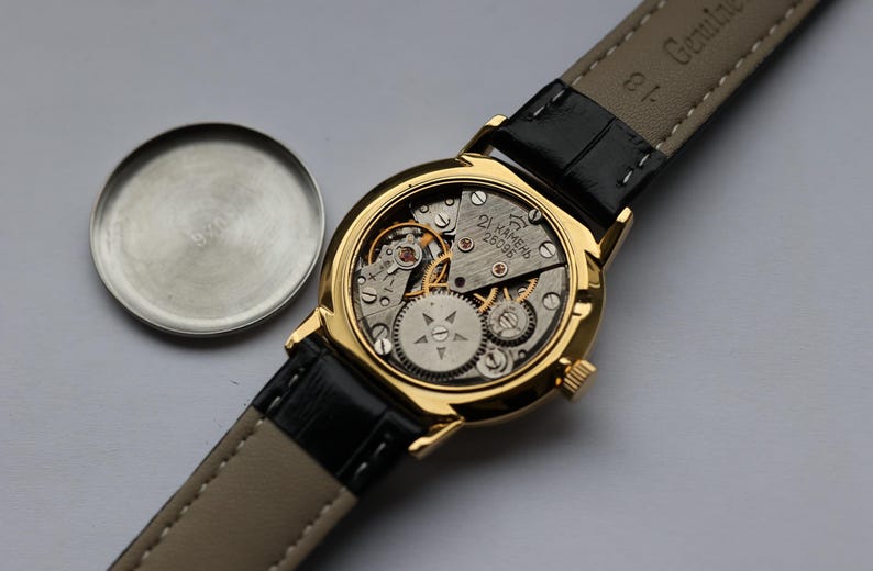 May include: A gold-toned wristwatch with a black leather strap, showcasing the intricate internal mechanisms. The watch face is open, revealing gears and components. The watch's back cover is detached and placed to the side.