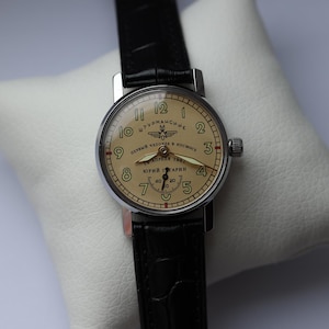 May include: A vintage wristwatch with a cream-colored face and black leather band. The watch face features black numerals, a seconds dial, and Cyrillic text. The watch is displayed on a white pillow.