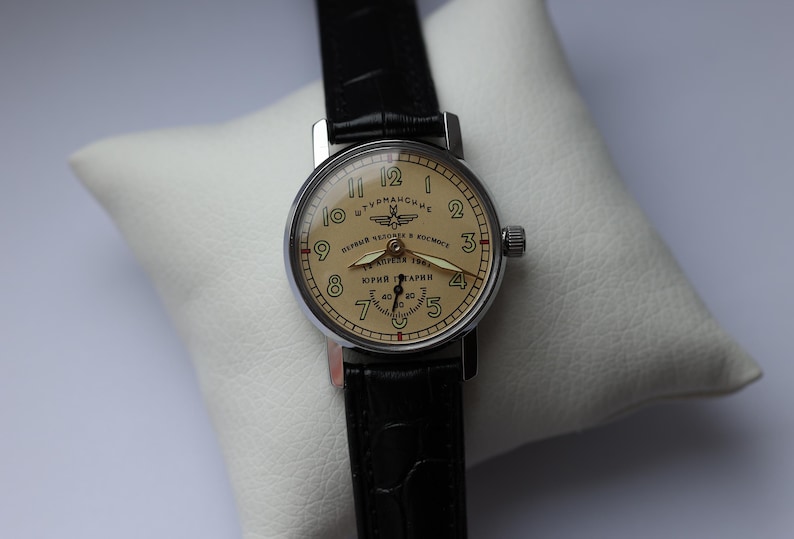 May include: A vintage wristwatch with a cream-colored face and black leather band. The watch face features black numerals, a seconds dial, and Cyrillic text. The watch is displayed on a white pillow.