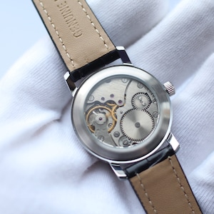 May include: A silver watch with a black leather band. The back of the watch is clear, revealing the intricate inner workings of the movement.