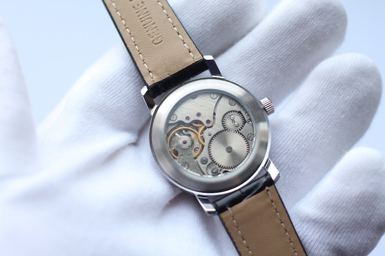 May include: A silver watch with a black leather band. The back of the watch is clear, revealing the intricate inner workings of the movement.