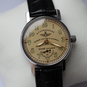 May include: A vintage wristwatch with a cream-colored face and black leather band. The watch face features black numerals, hands, and text in Cyrillic script. The watch is a Sturmanskie model, commemorating Yuri Gagarin's spaceflight.