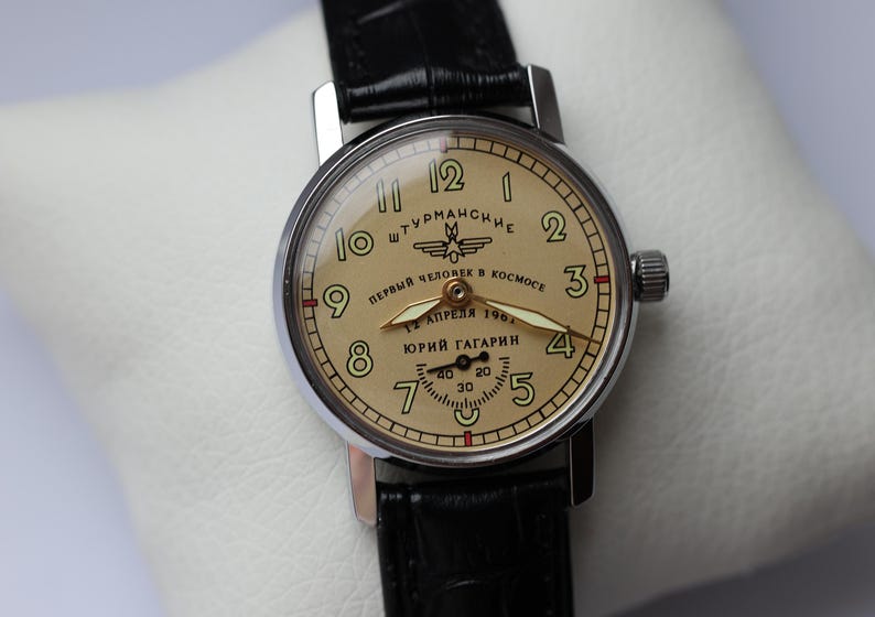 May include: A vintage wristwatch with a cream-colored face and black leather band. The watch face features black numerals, hands, and text in Cyrillic script. The watch is a Sturmanskie model, commemorating Yuri Gagarin's spaceflight.