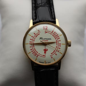 May include: Vintage gold-toned wristwatch with a cream-colored face and black leather band. The watch face features red numerals and text, including the word "Baltika". The watch has a gold-toned crown and a black leather strap.