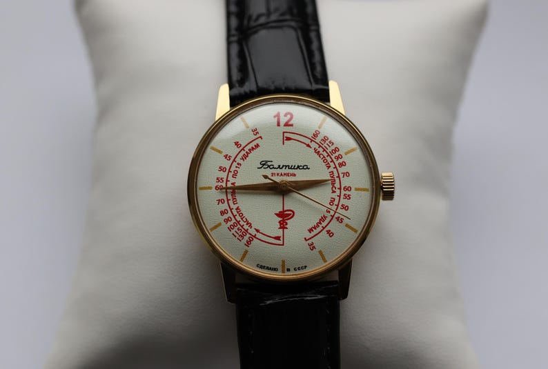 May include: Vintage gold-toned wristwatch with a cream-colored face and black leather band. The watch face features red numerals and text, including the word "Baltika". The watch has a gold-toned crown and a black leather strap.