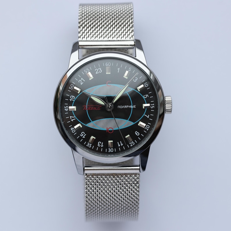 May include: A silver-toned wristwatch with a black face featuring a unique design with blue and red accents. The watch has a mesh band and a dial with numbers and text, including the word "Polarnye".